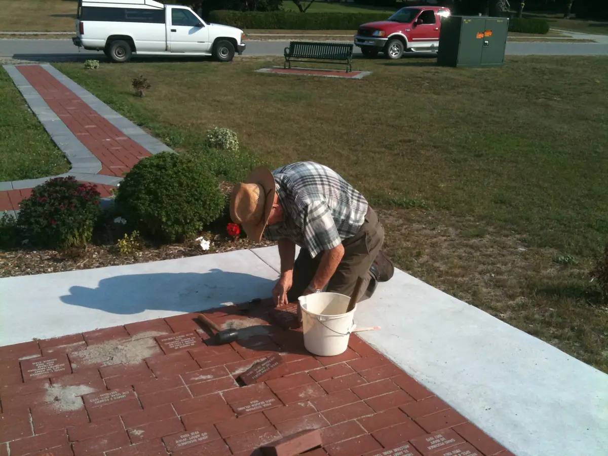 A man kneels on a brick-paved memorial area while working beside inscribed plaques, tools, and a bucket.