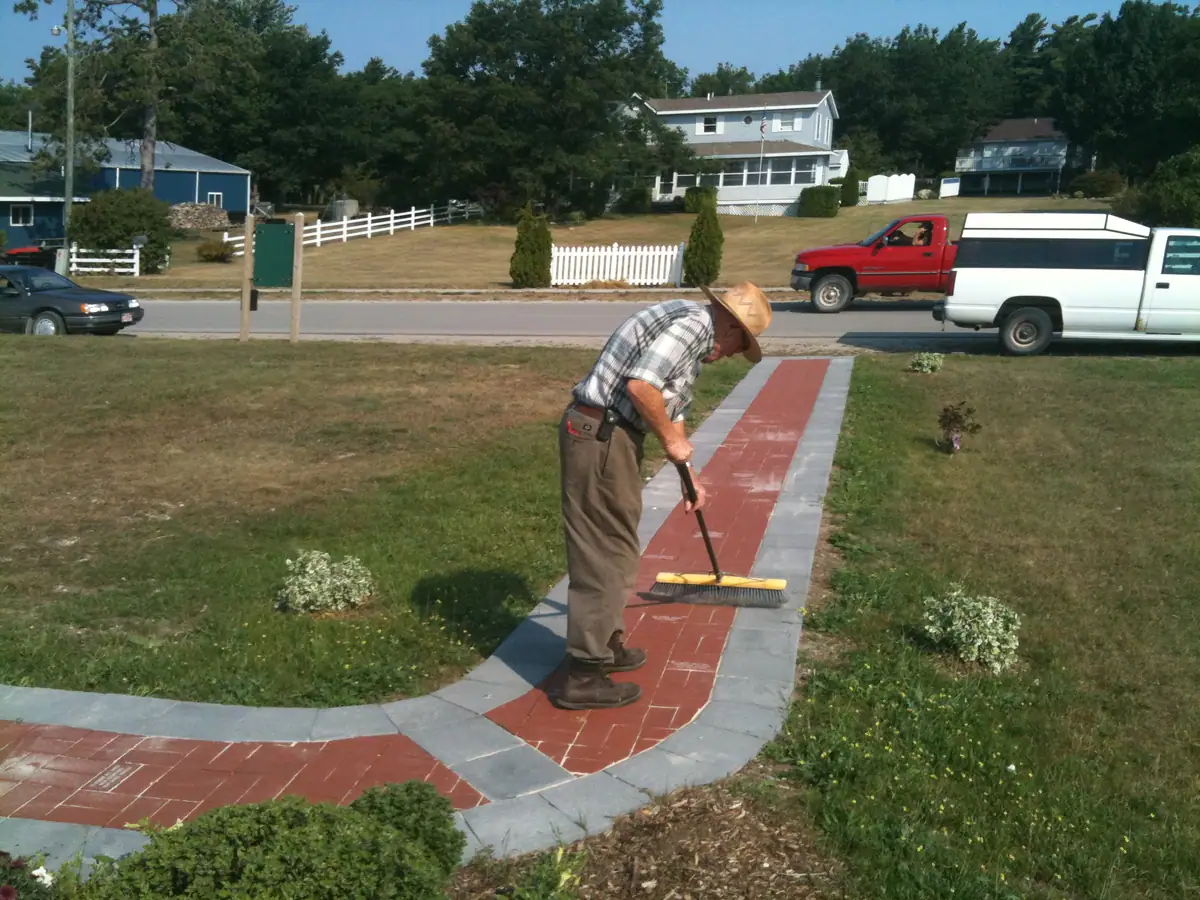 A man in a straw hat sweeps a red brick walkway beside a residential street.