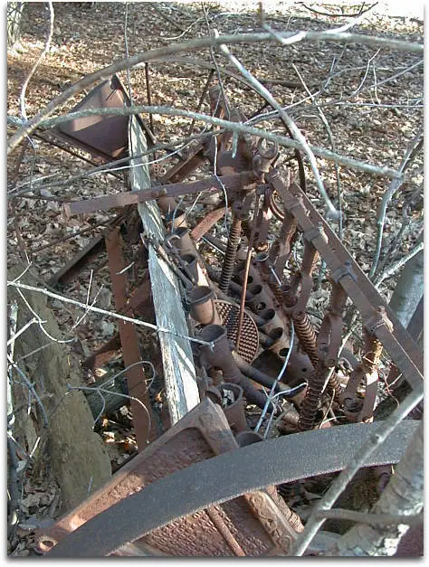 A rusted farm machine with springs and metal frames lies among trees and leaf litter.