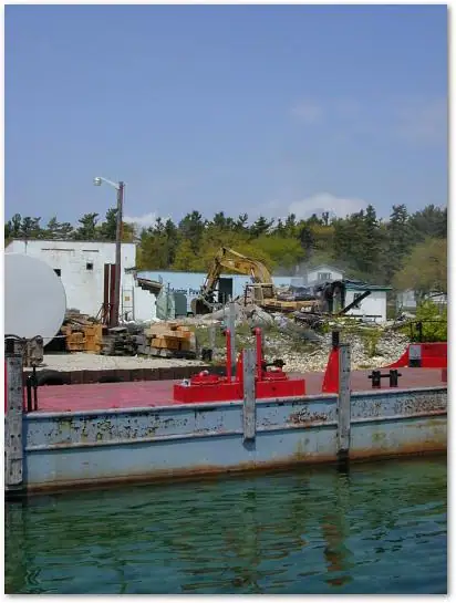 A blue-and-white barge sits in front of a waterfront demolition site with an excavator and rubble.
