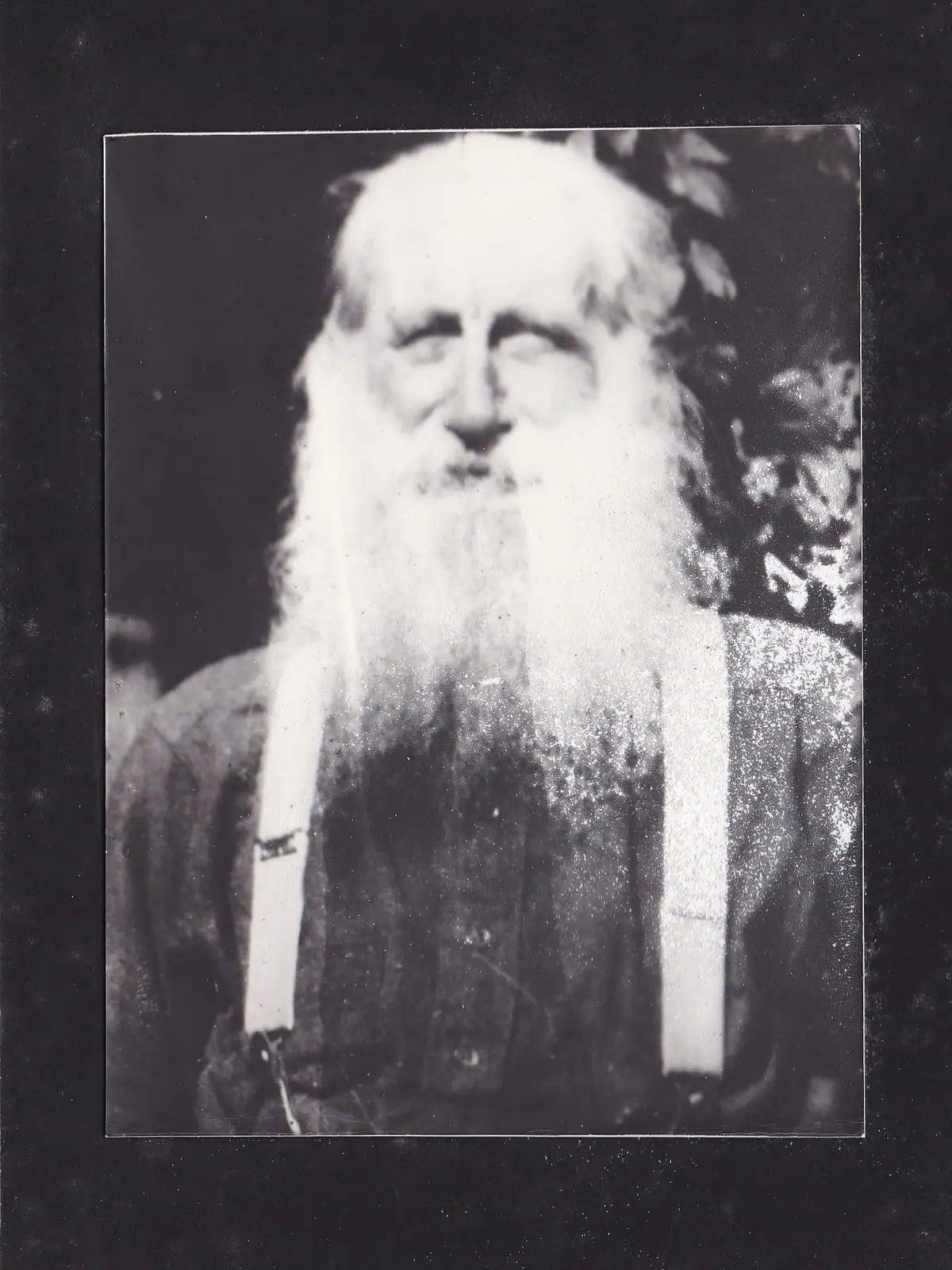 Black-and-white portrait of an older man with a long white beard, dark clothing, and light suspenders.