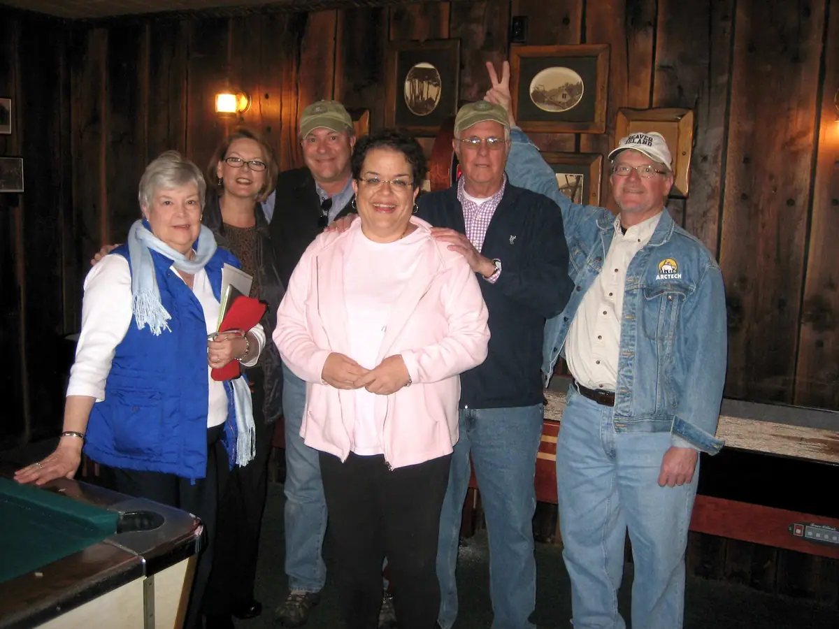 Six adults pose in a wood-paneled room beside a pool table, with a partially hidden person making a peace sign.