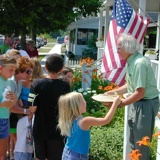 Children and adults gather beside a flower-filled porch as a girl receives a woven basket beneath an American flag.