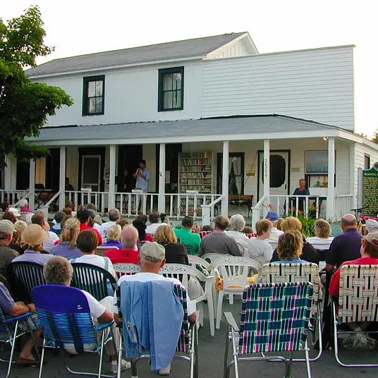 A seated outdoor audience listens to a speaker on the porch of a white-sided building.