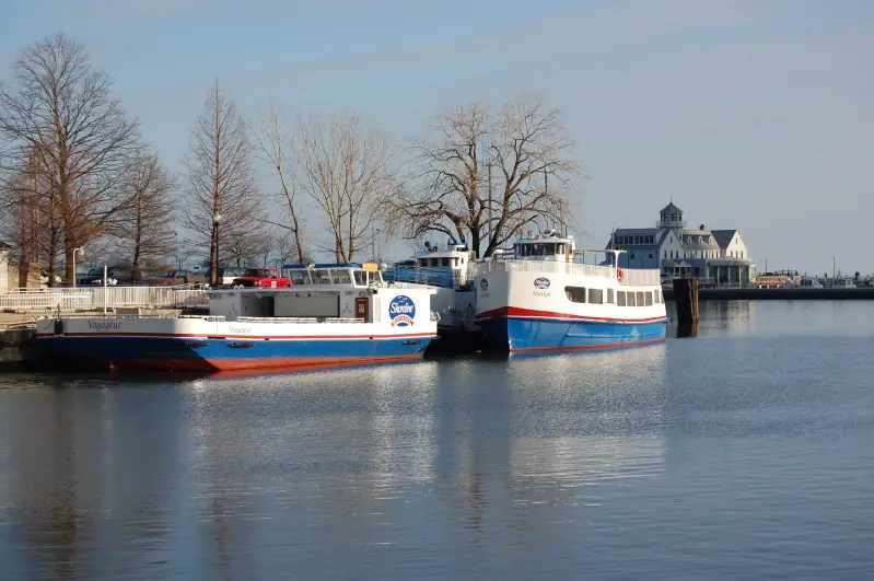 Two passenger ferries are moored beside a pier on calm water, with leafless trees and a gabled waterfront building behind them.