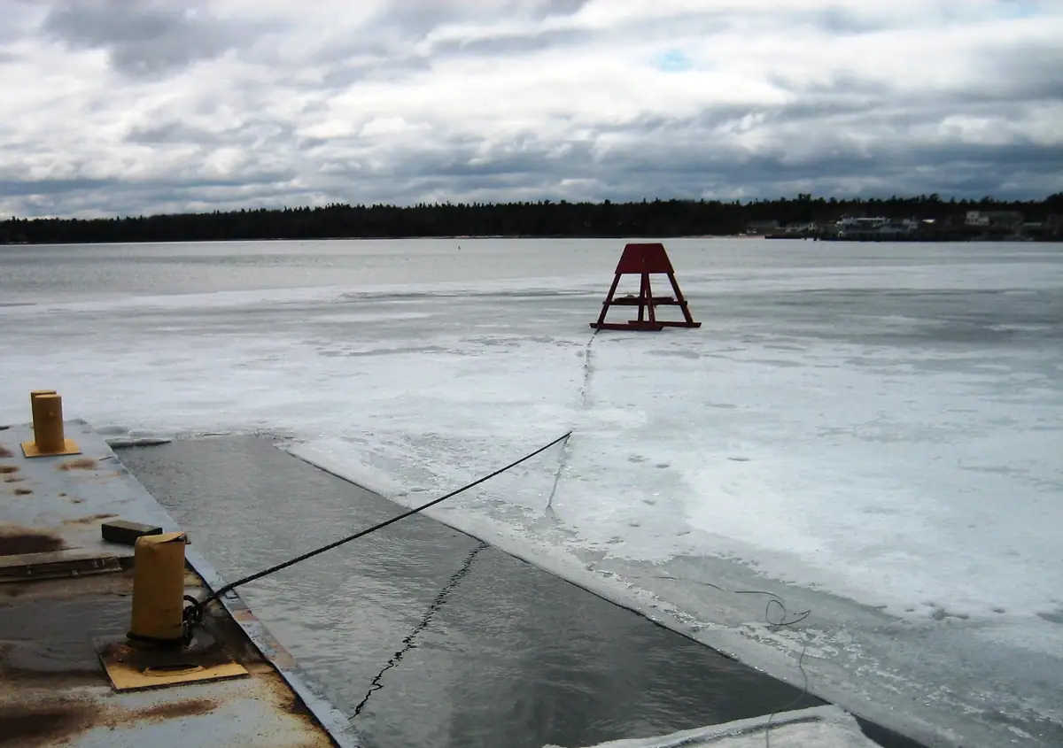 A dock, mooring rope, and red marker stand beside ice-covered water under a cloudy sky.