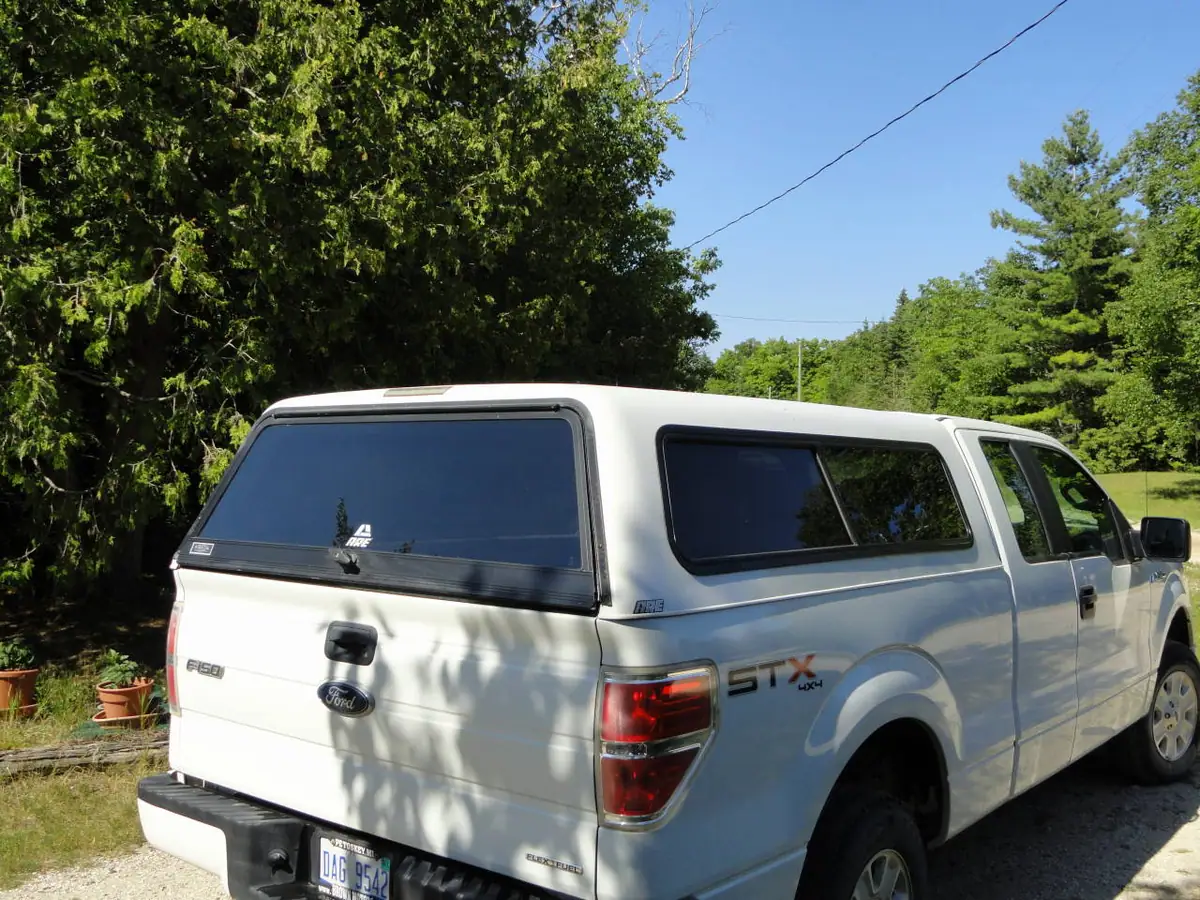 White pickup truck with a camper shell parked beside trees on a sunny gravel drive.