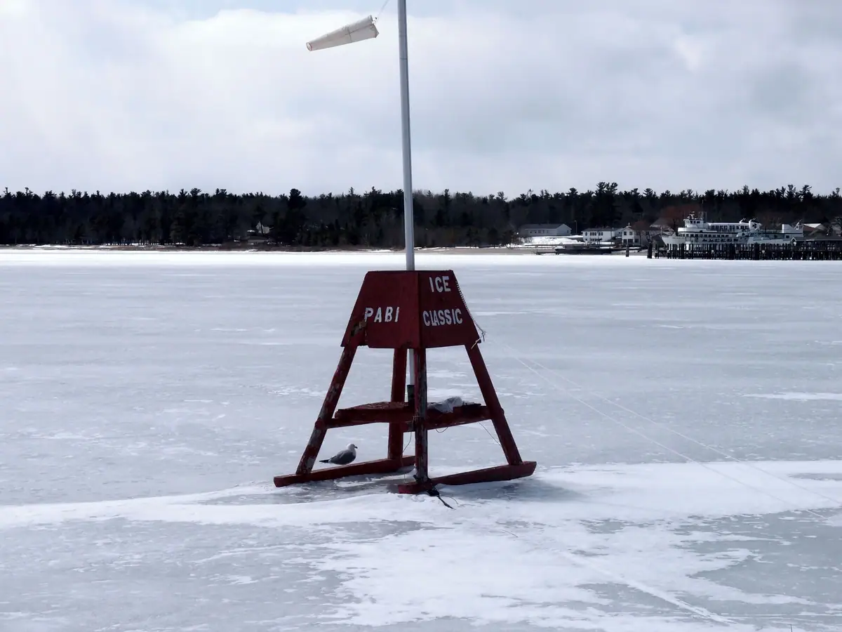 A red A-frame marker stands on frozen water with a gull, windsock, dock, and ferry in the distance.