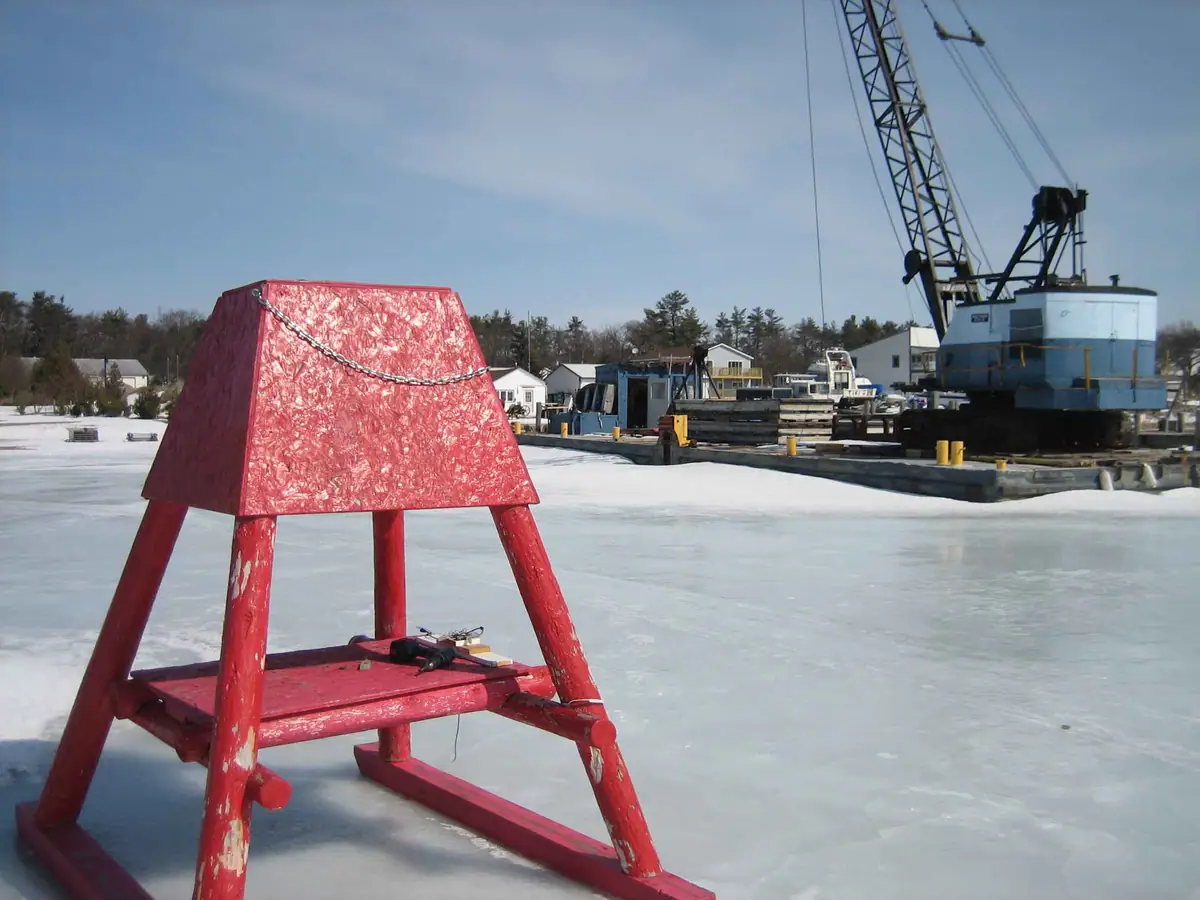 A red wooden structure stands on ice beside a dock and large construction crane.