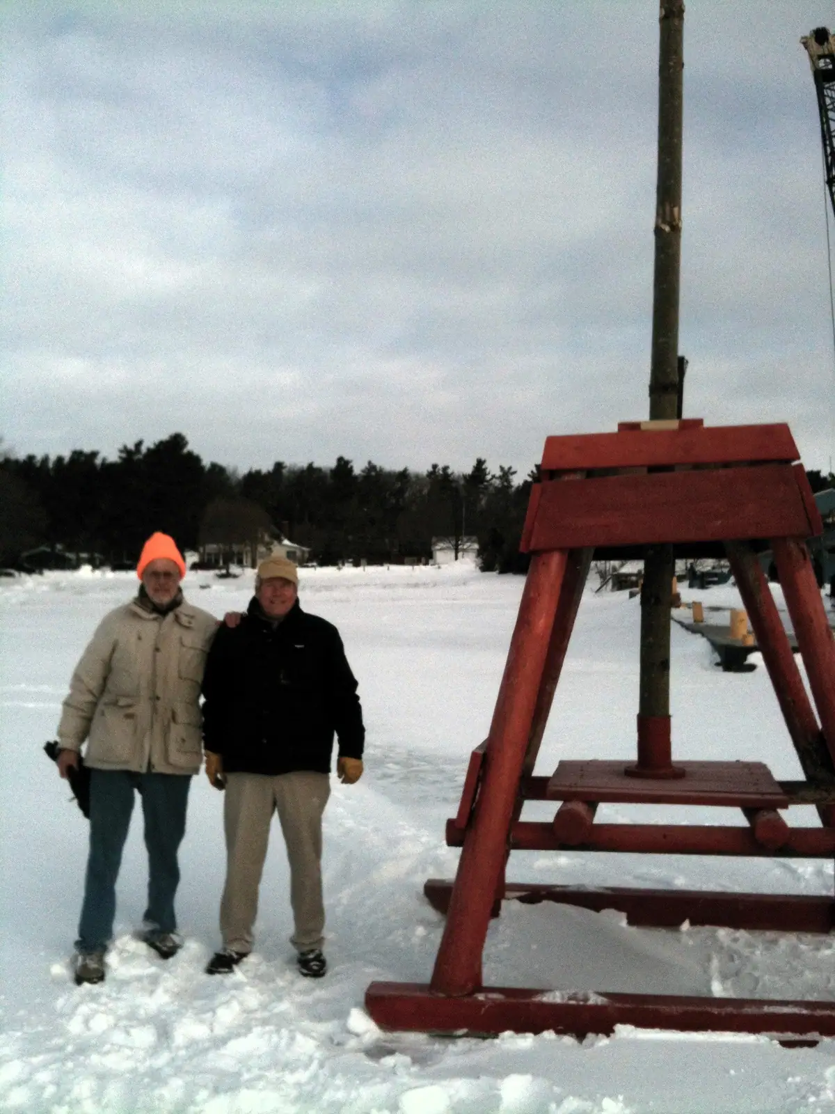 Two men stand beside a red wooden frame on a snow-covered expanse in winter.
