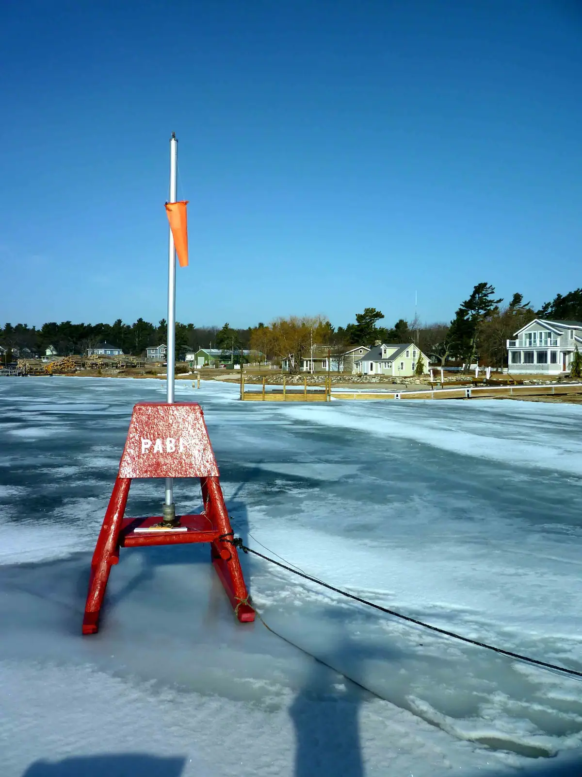 A red A-frame marker with an orange flag stands on frozen water near houses and a wooden dock.