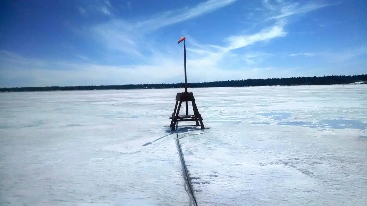 A windsock mounted on a wooden tripod stands on a broad frozen expanse beneath a blue sky.