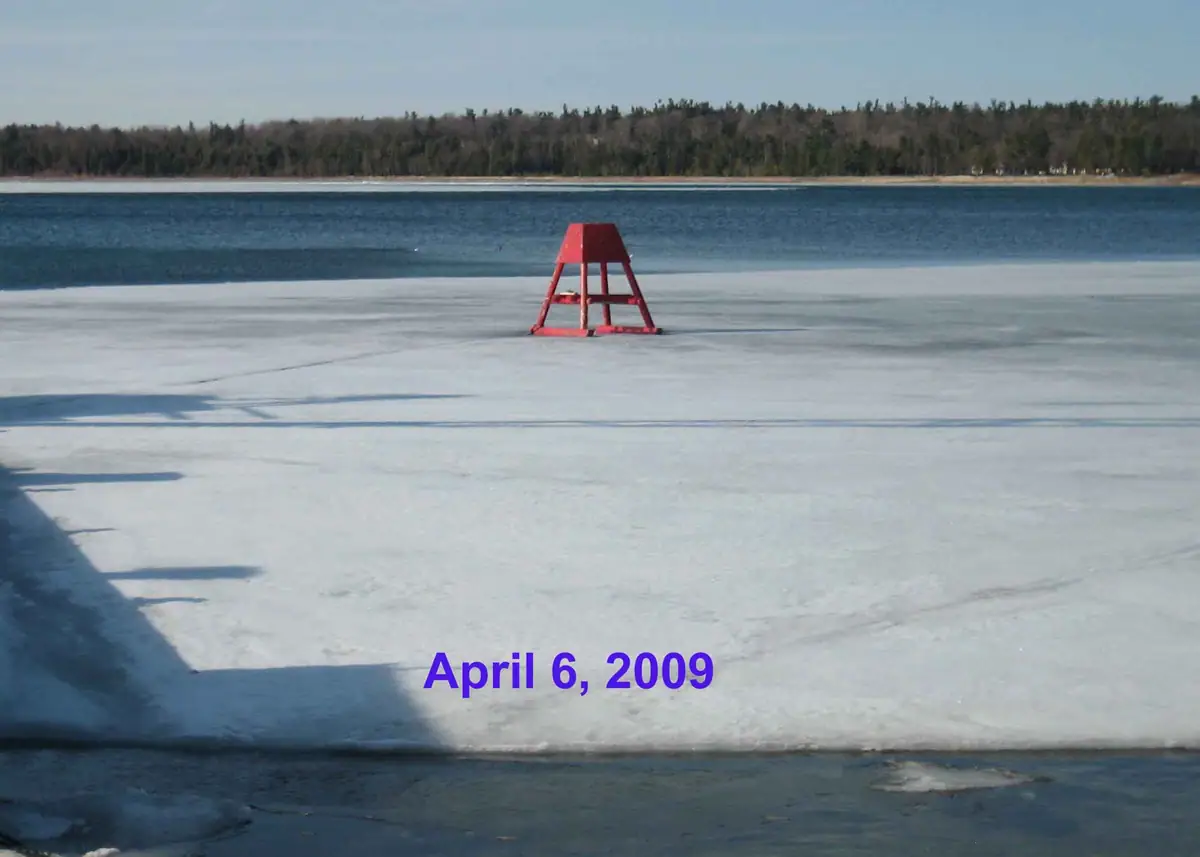 A red navigation marker stands on ice beside open water and a forested shoreline.