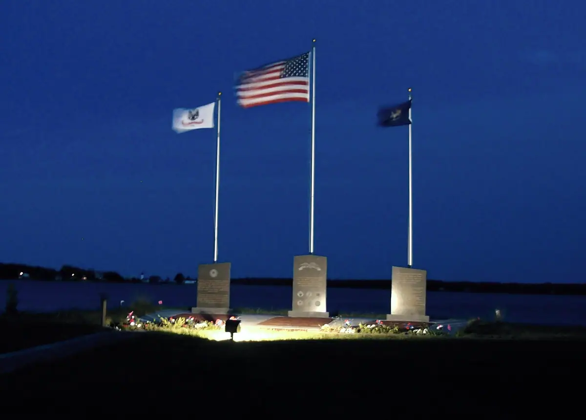 Illuminated nighttime memorial with three flagpoles, stone plaques, flowers, and water behind it.