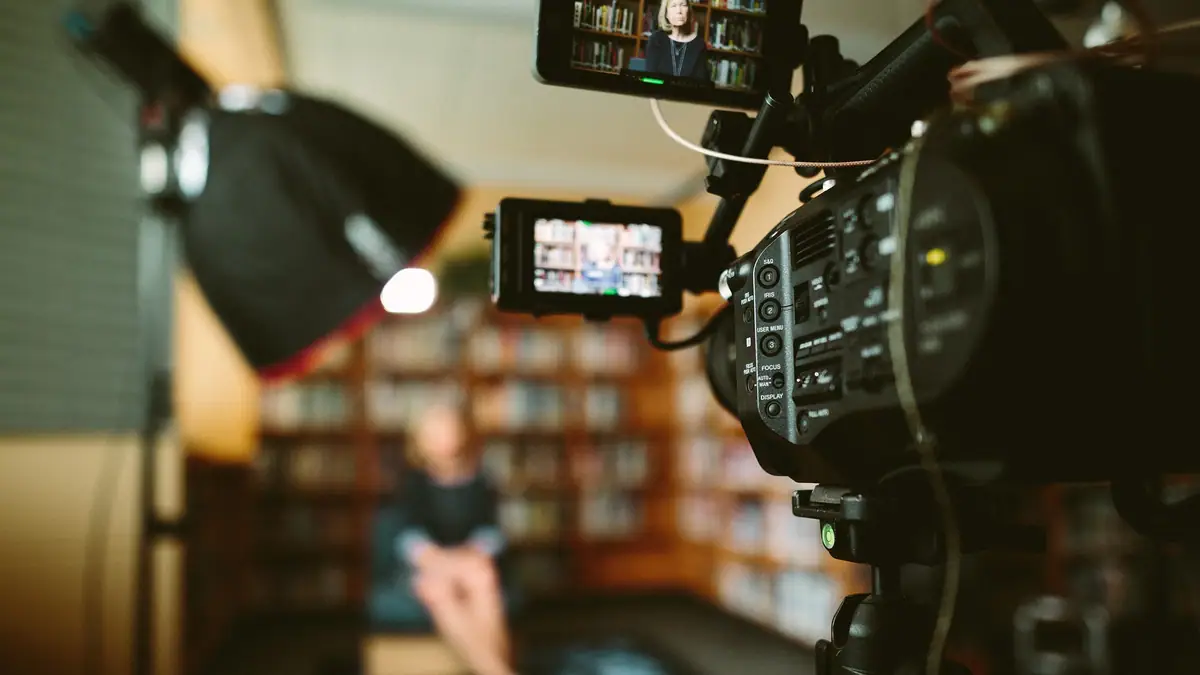 A professional video camera records a seated person in a room lined with bookshelves.