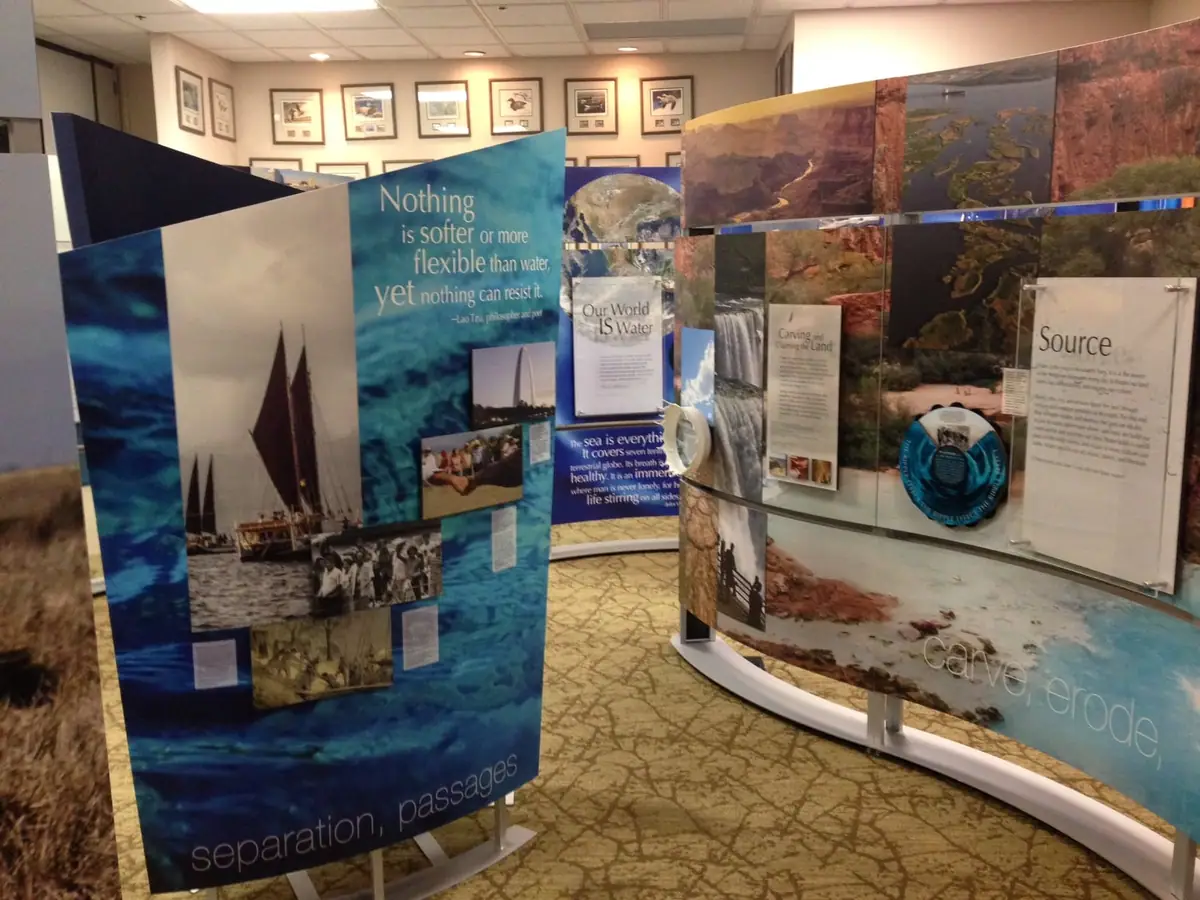 Indoor exhibition display with blue panels, water-themed photographs, landscape images, and printed interpretive text.
